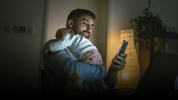 Smiling man holding baby whilst looking at phone in dimly lit room