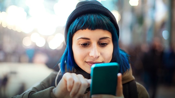 A young woman with bright blue hair underneath a beanie hat taps on her phone.