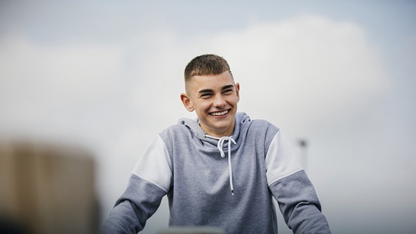 A teenage boy in a grey hoodie looks at the camera and smiles.