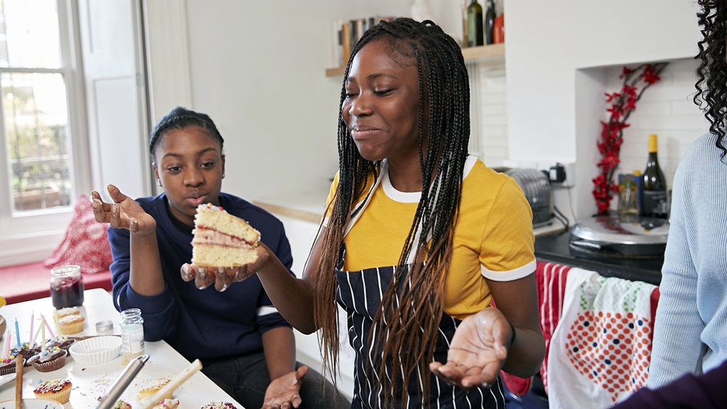 Two friends in aprons smile as they hold slices of the cake they've just baked