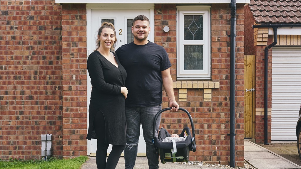New parents stood in front garden holding baby in carrier