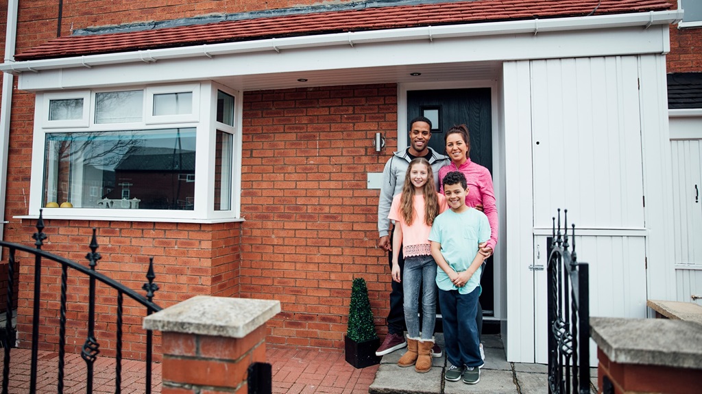 Family stands on the front doorstep outside their home smiling to the camera