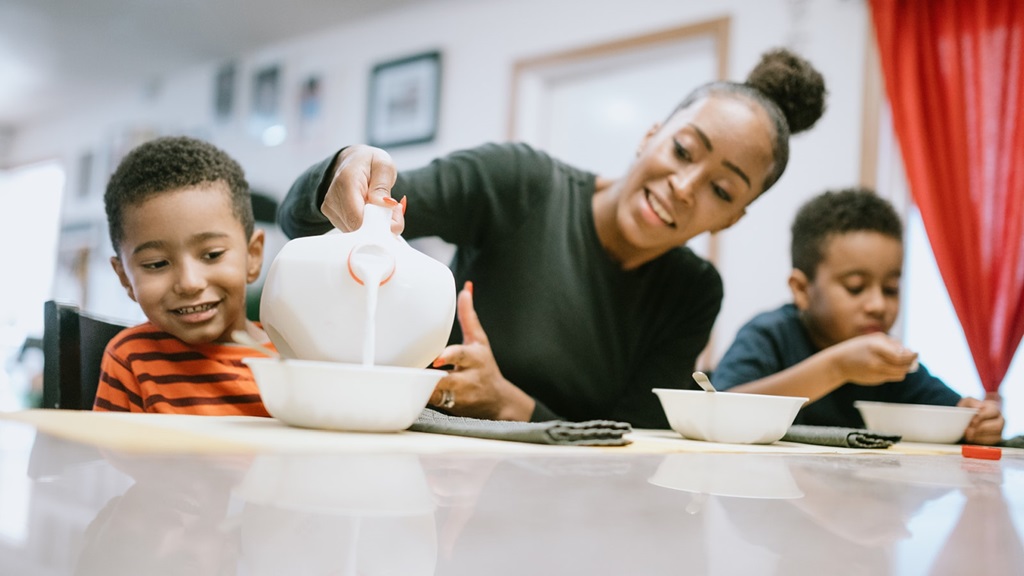 Woman with two children sat at table pours milk into a cereal bowl 