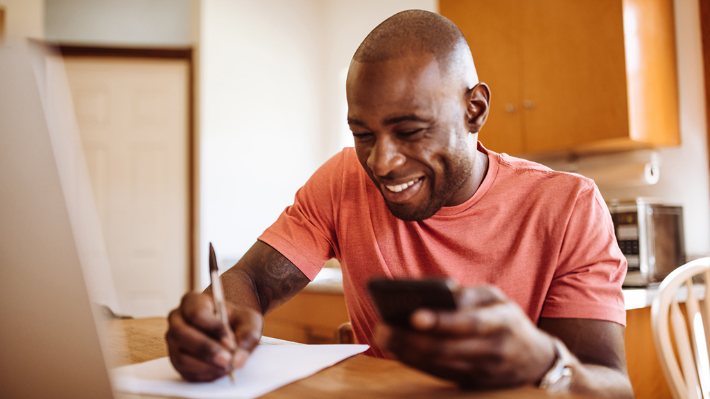 Man at kitchen table smiling and writing whilst looking at his phone