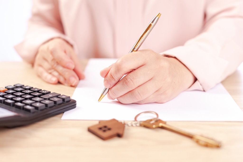 A person's hands signing a document, with house keys and a calculator sitting on the desk next to them