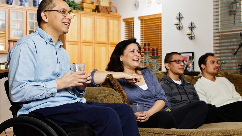 Two parents sit with their adult sons on a sofa, all smiling while watching the television