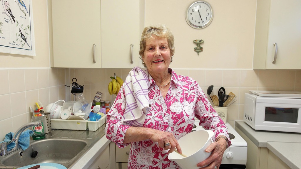 Florence in her kitchen holding a mixing bowl, behind her is an oven, to the left a sink and draining board. She is a resident.