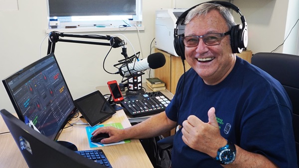 Steve Medlin sits at a desk with two laptops, a microphone and recording equipment. He's smiling and giving the thumbs up