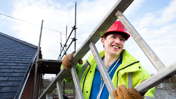 Man in hard hat and high-vis smiling while carrying a ladder. Scaffolding surrounds the house roof next door.